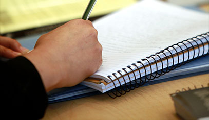 Close up image someone's hand. The hand is holding a pen and writing in a spiral notebook.